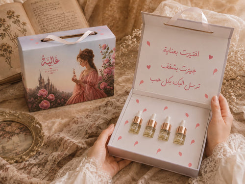 A person holds an open white gift box containing four small cosmetic dropper bottles with rose-gold caps. Beside it is the closed matching box, decorated with a vintage-style illustration of a woman in a pink dress and pink roses, with Arabic text on the inside lid.